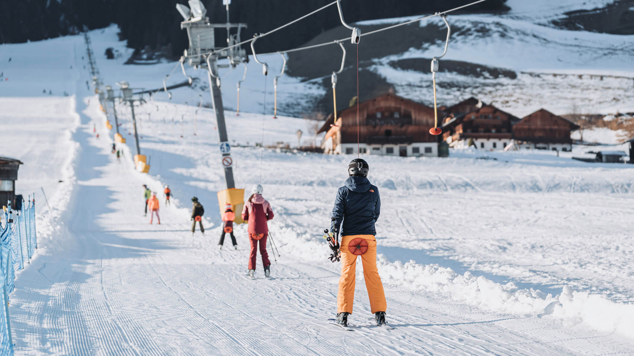 Skifahrer, die mit einem Skilift fahren und auf einer verschneiten Piste in der Nähe von Holzhütten in einem Gebirgsort stehen. - Hotel Magdalenahof