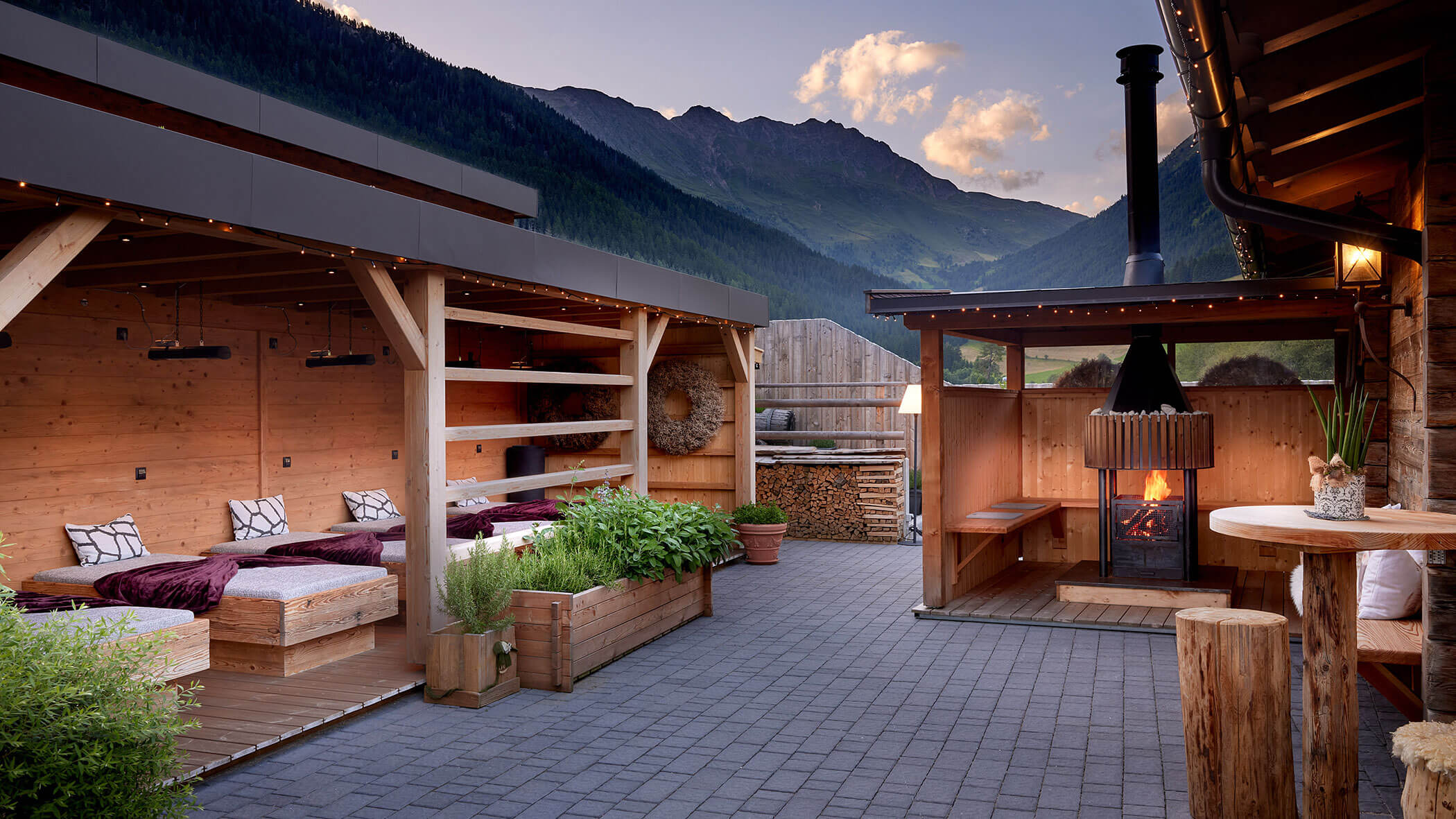 Gemütliche Außenterrasse mit Sitzgelegenheiten aus Holz, Kaminofen und Bergblick bei Sonnenuntergang. - Hotel Magdalenahof