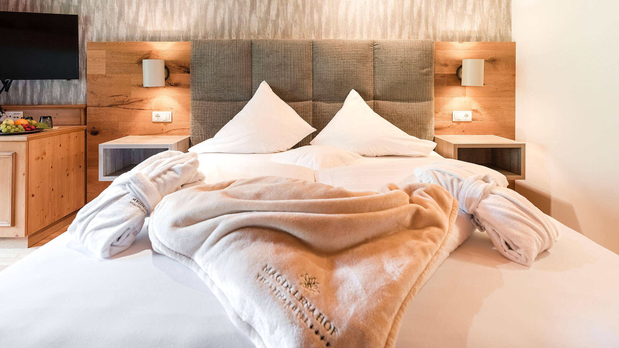 Hotelzimmer mit Doppelbett, flauschigen Bademänteln, Holzakzenten und einem Fernseher auf einem Schrank. - Hotel Magdalenahof