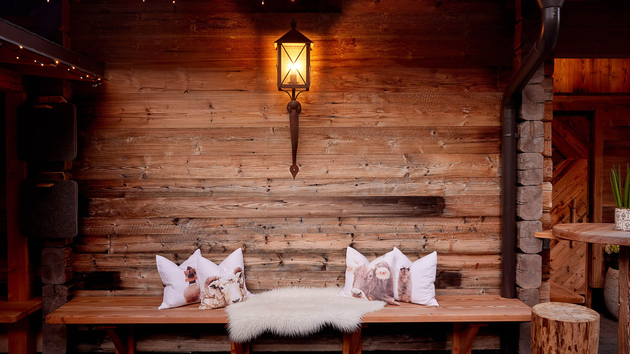 Holzbank mit Kissen mit Tiermotiven und einer weißen Felldecke vor einer rustikalen Holzwand mit einer Laterne. - Hotel Magdalenahof