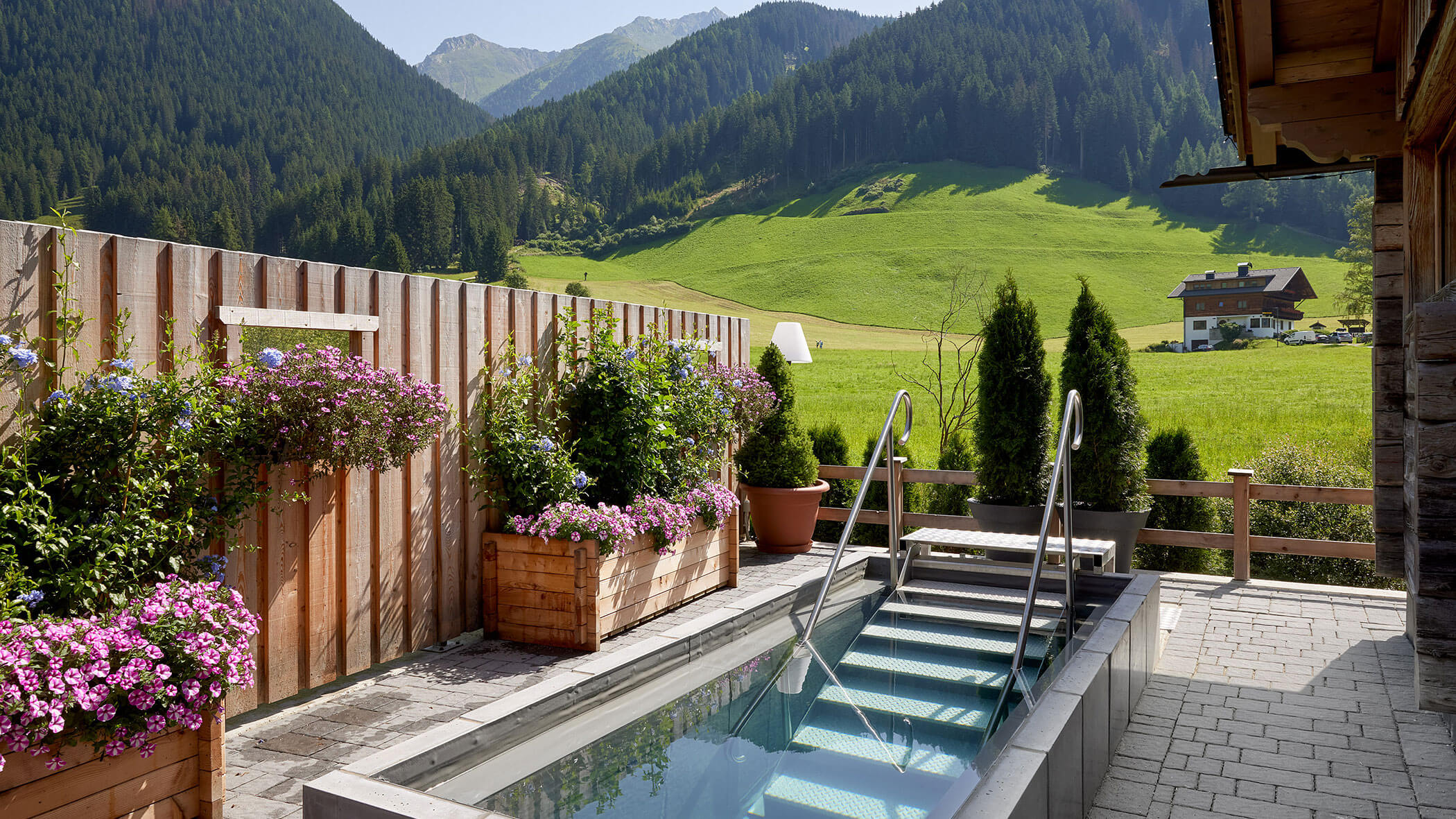 Ein kleiner Pool mit Stufen, umgeben von Blumen, blickt auf grüne Hügel und Berge unter einem klaren Himmel. - Hotel Magdalenahof