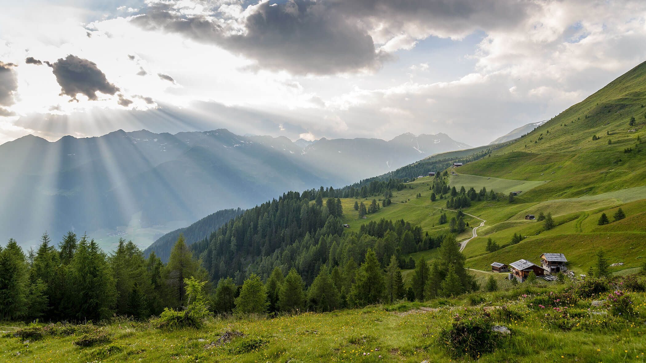 Sonnenstrahlen scheinen durch die Wolken über grüne Hügel, Wälder und verstreute Häuser in einer Berglandschaft. - Hotel Magdalenahof