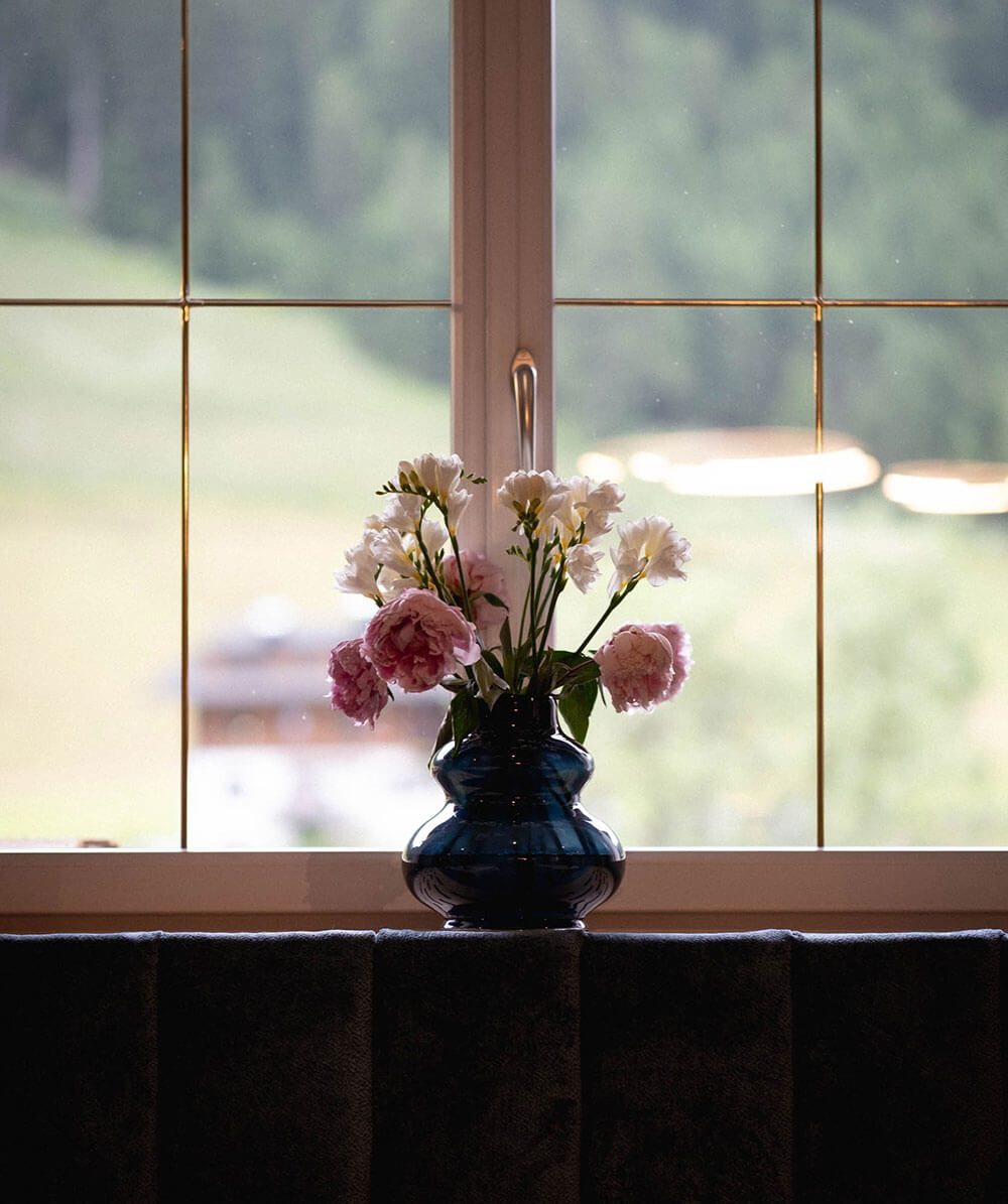 Eine dunkle Vase mit rosa und weißen Blumen steht auf einer Fensterbank vor einem hellen Fenster. - Hotel Magdalenahof