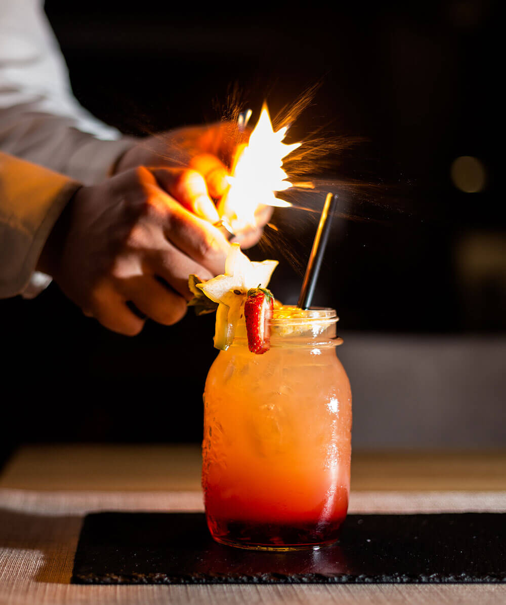 Eine Person entzündet eine Flamme über einem Fruchtcocktail in einem Einmachglas, garniert mit Früchten und einem Strohhalm. - Hotel Magdalenahof