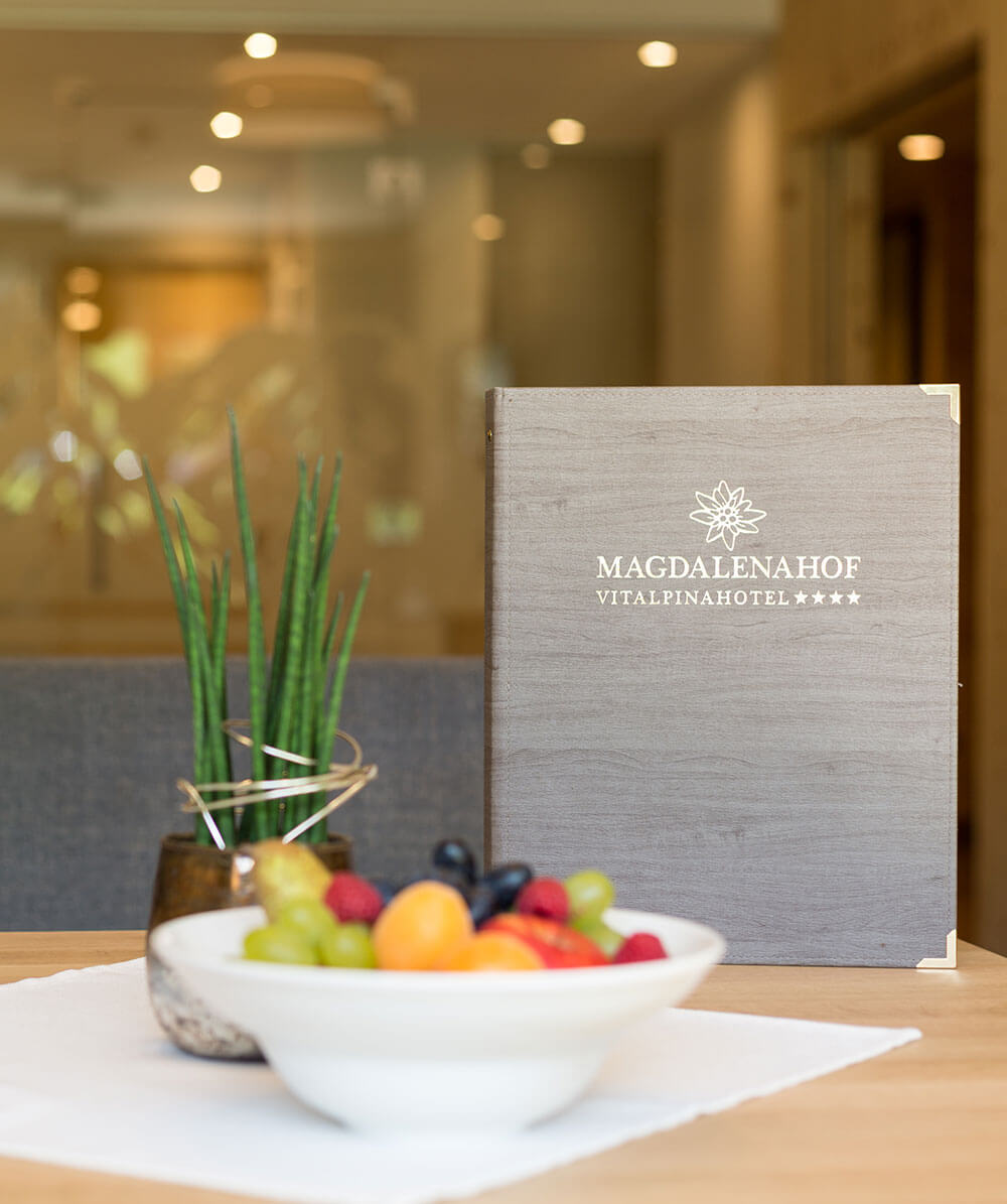 Eine Schale mit frischem Obst und eine Topfpflanze auf einem Tisch neben einer Hotelkarte mit der Aufschrift Magdalenahof. - Hotel Magdalenahof