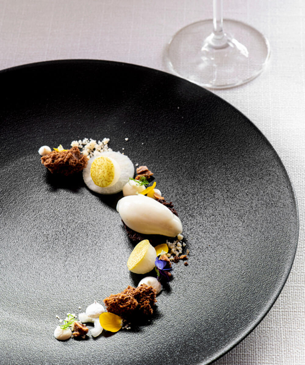 Kunstvoll angerichtetes Dessert mit Sahnetupfen, Biskuitstückchen und Quenelle auf einem schwarzen Teller; daneben ein Weinglas. - Hotel Magdalenahof