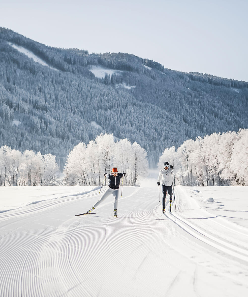 Zwei Personen beim Skilanglauf auf einer verschneiten Loipe mit schneebedeckten Bäumen und Bergen im Hintergrund. - Hotel Magdalenahof