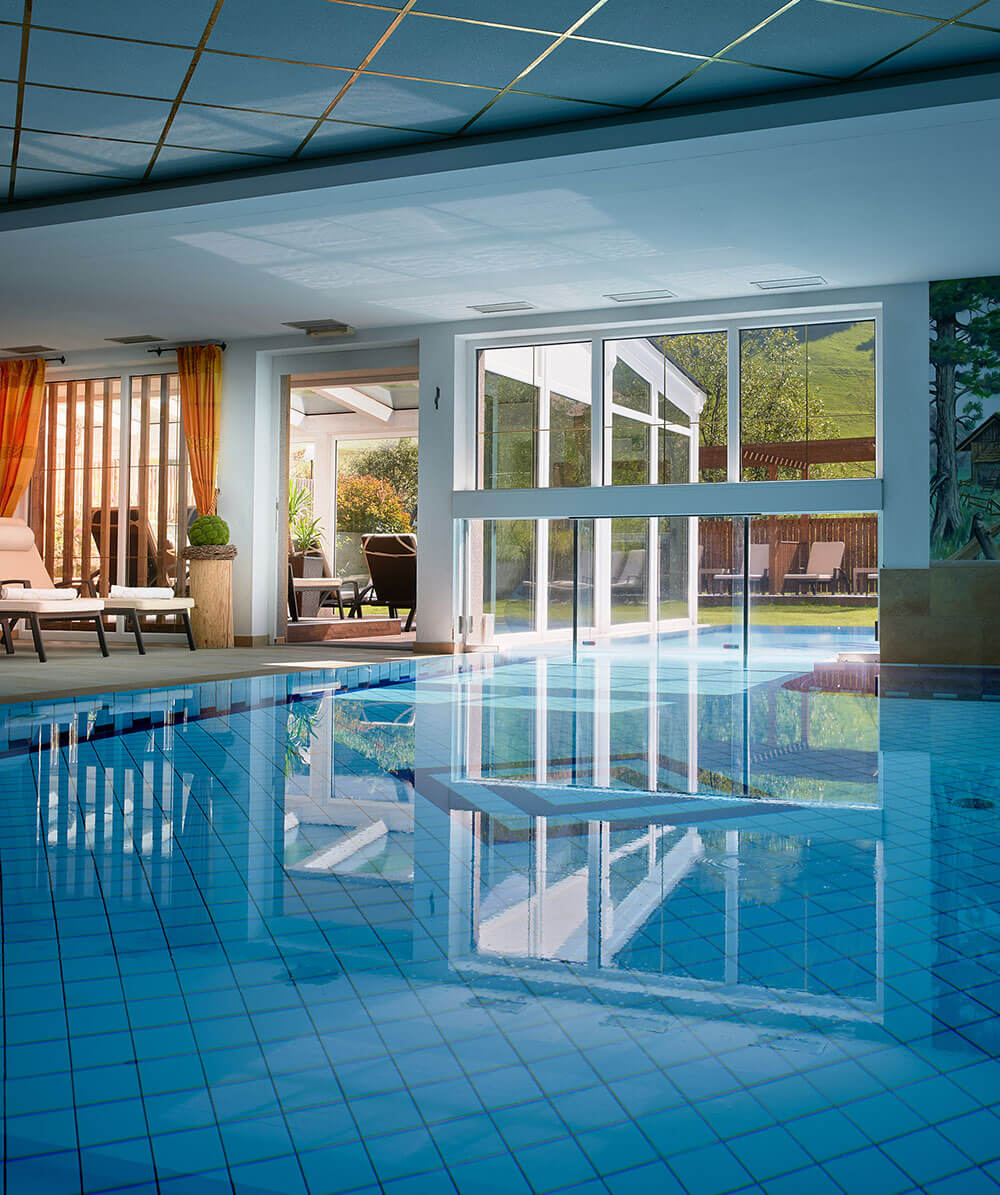 Innenpool mit blauen Fliesen, großen Fenstern und Liegestühlen; Sonnenlicht strömt durch offene Glastüren. - Hotel Magdalenahof