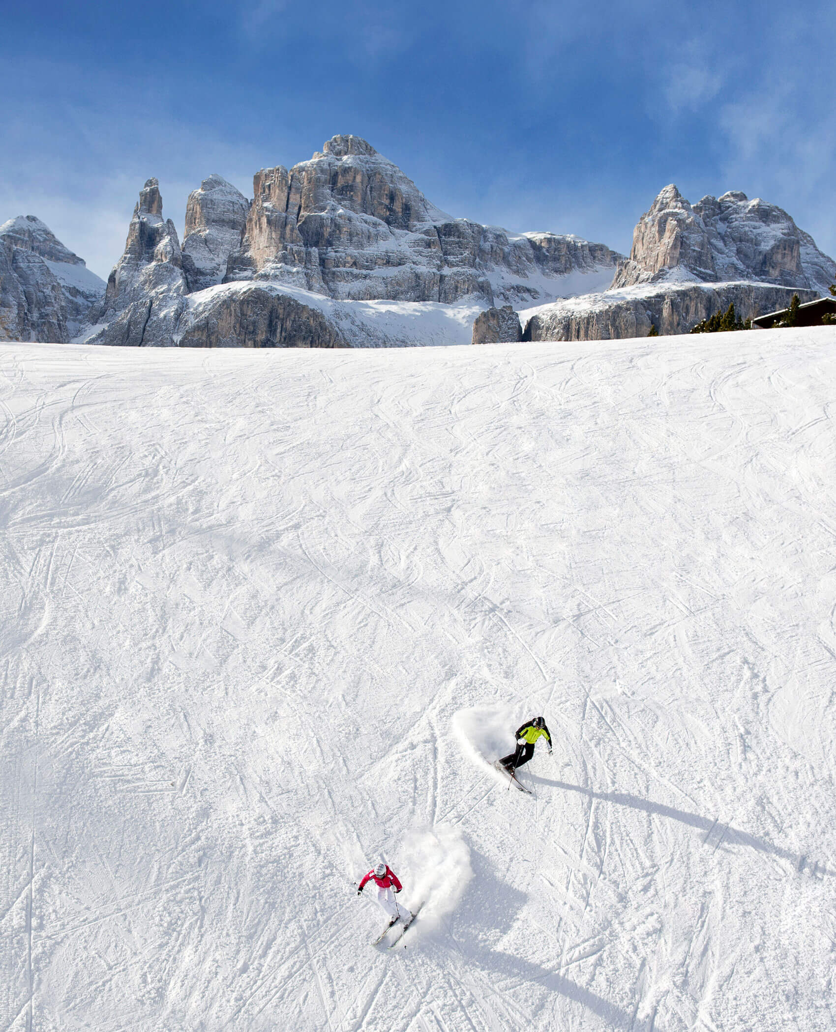 Zwei Skifahrer carven durch den Neuschnee auf einer weiten Piste mit felsigen Bergen im Hintergrund unter blauem Himmel. - Hotel Magdalenahof