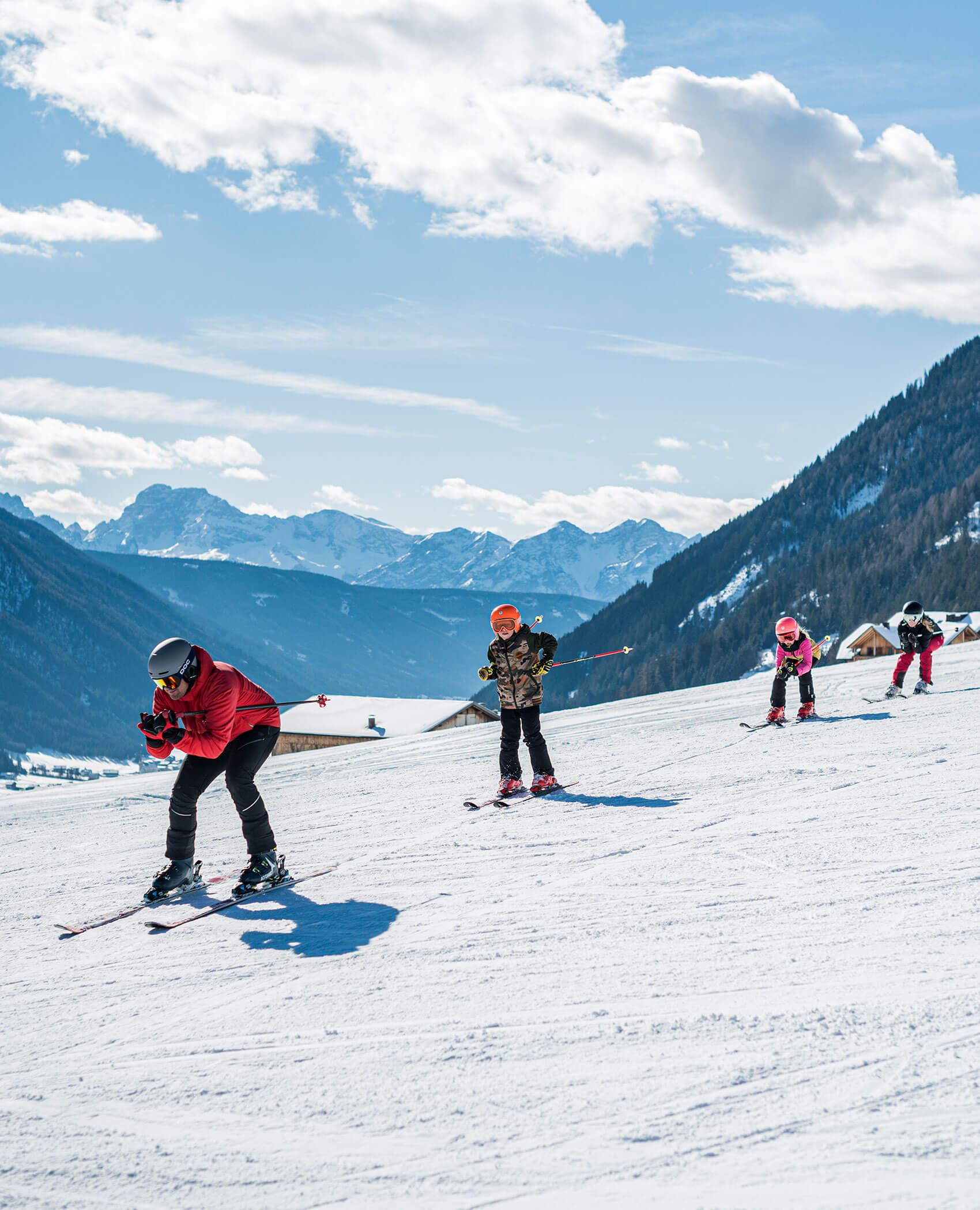 Vier Personen fahren auf einer verschneiten Piste mit Bergen und blauem Himmel im Hintergrund. - Hotel Magdalenahof