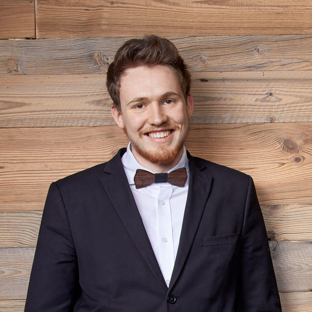 Mann im Anzug und mit Fliege, der lächelnd vor einer Bretterwand steht. - Hotel Magdalenahof