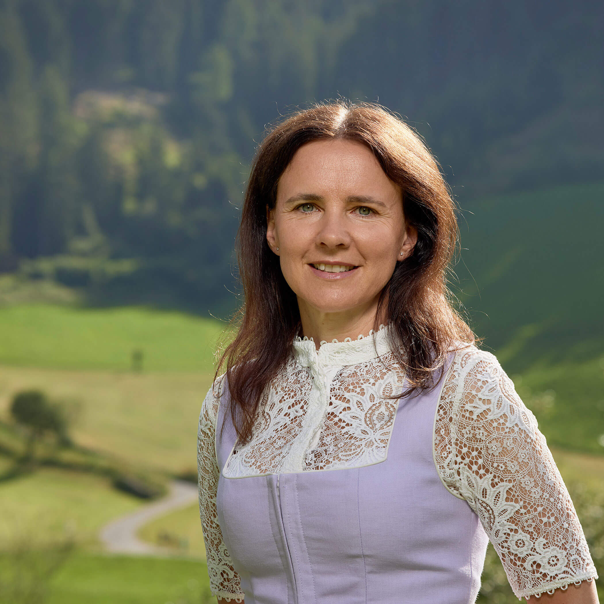 Frau mit langen braunen Haaren in einer Spitzenbluse steht im Freien mit grünen Hügeln und Bäumen im Hintergrund. - Hotel Magdalenahof