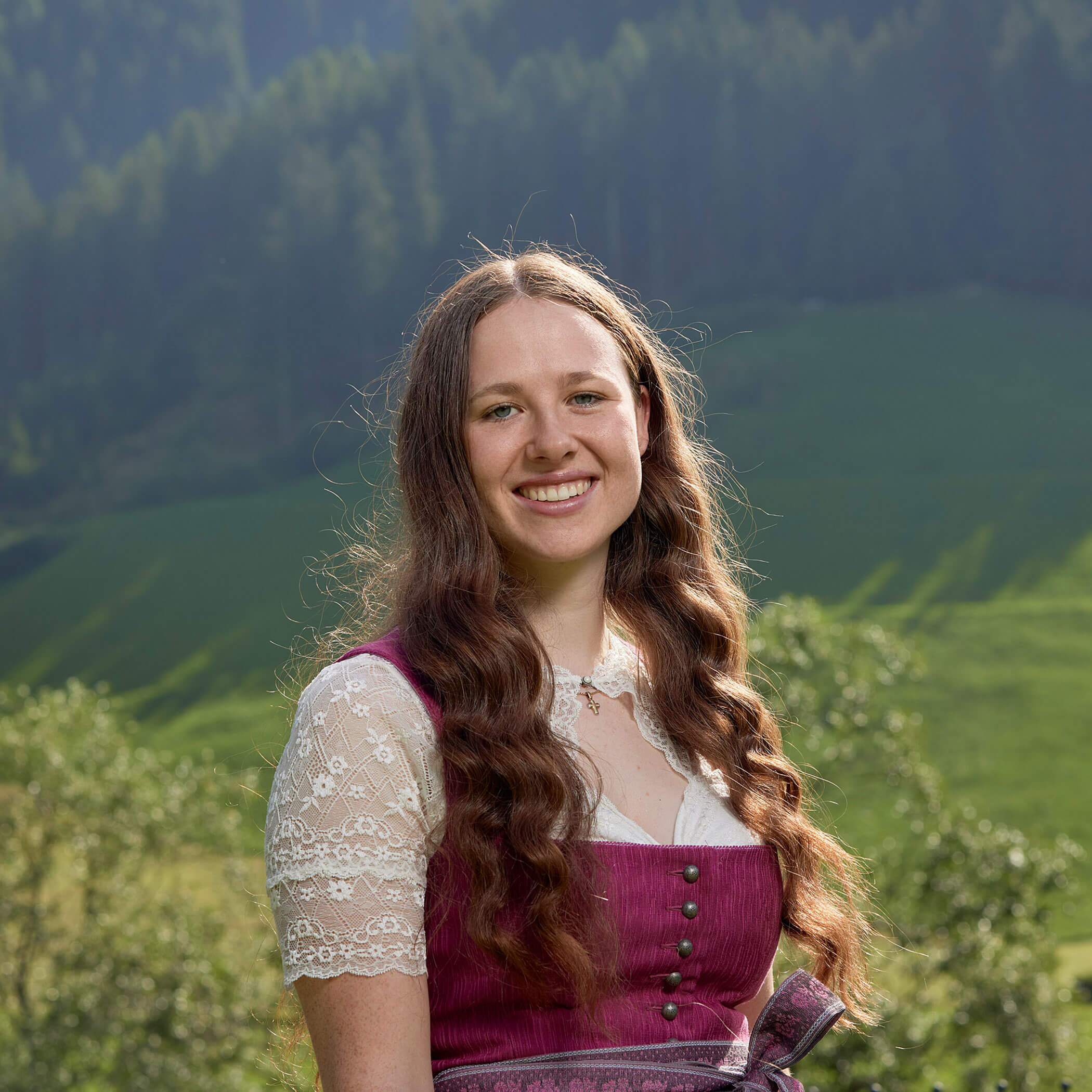 Eine junge Frau mit langem, gewelltem Haar lächelt im Freien, sie trägt ein traditionelles Kleid, im Hintergrund sind die Berge zu sehen. - Hotel Magdalenahof