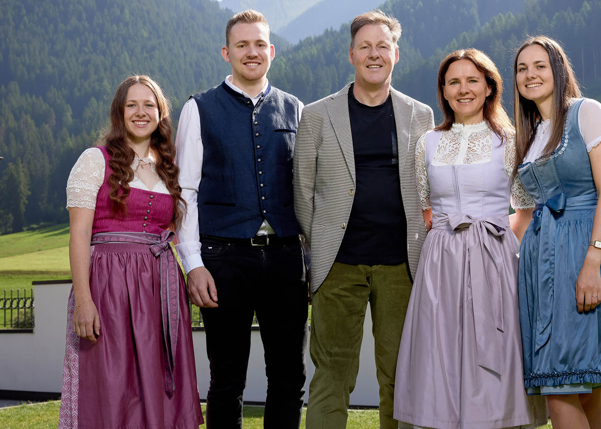 Five people in traditional Bavarian clothing stand smiling outdoors with mountains and greenery in the background. - Hotel Magdalenahof