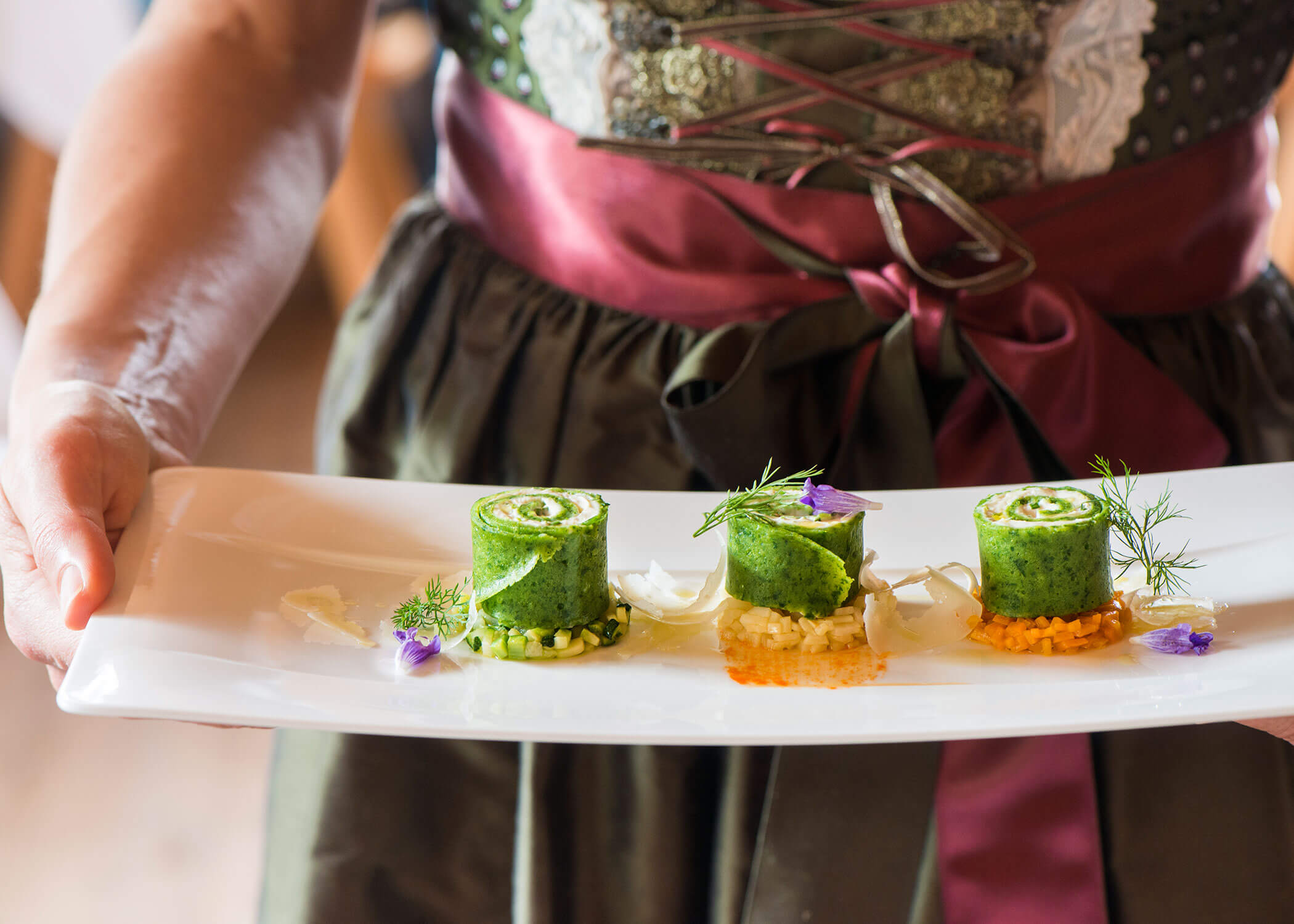 A person in traditional dress holds a plate with three rolled green appetizers, garnished with herbs and flowers. - Hotel Magdalenahof