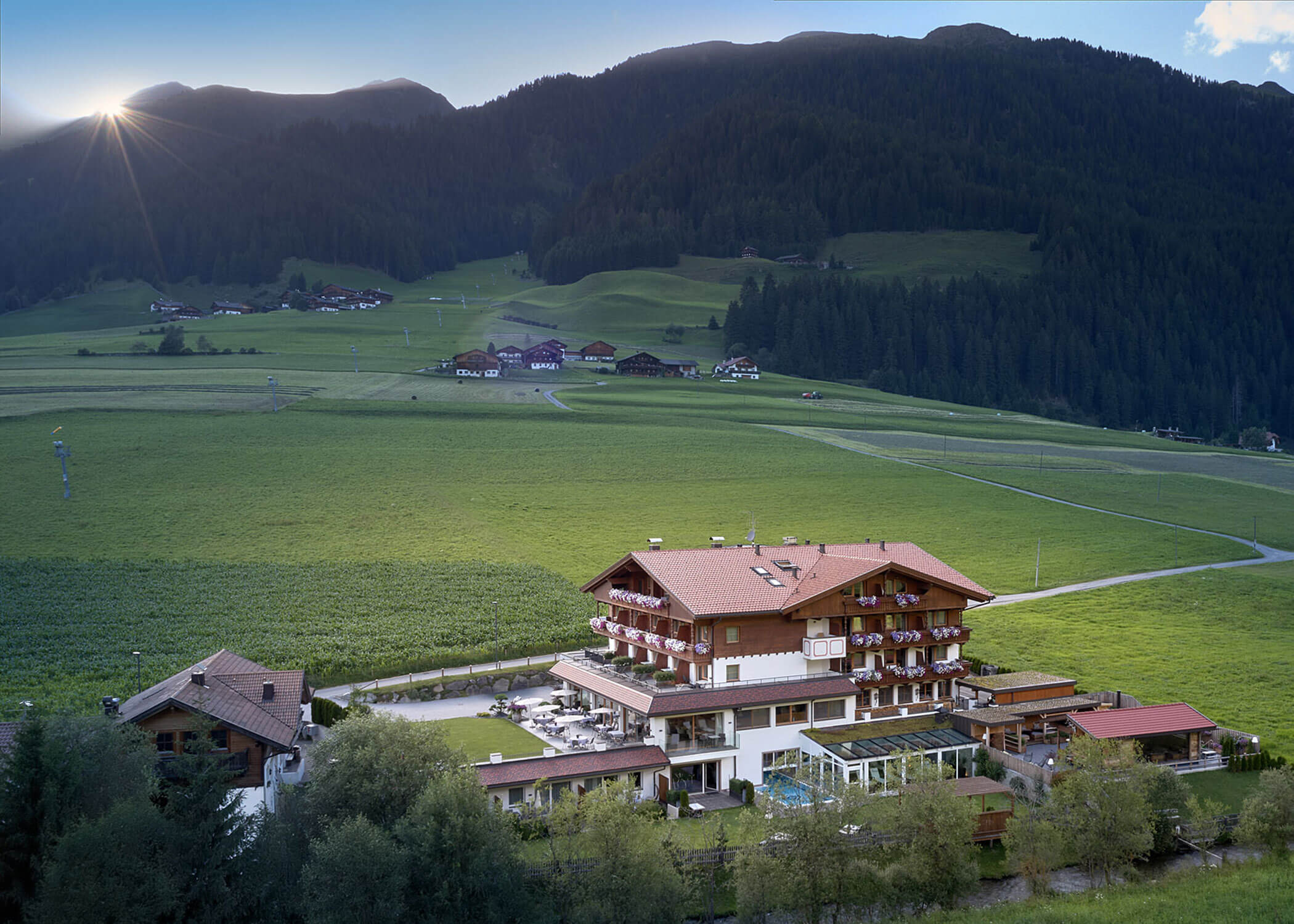 Alpine hotel with red roof in a green valley, surrounded by mountains and scattered houses at sunrise. - Hotel Magdalenahof
