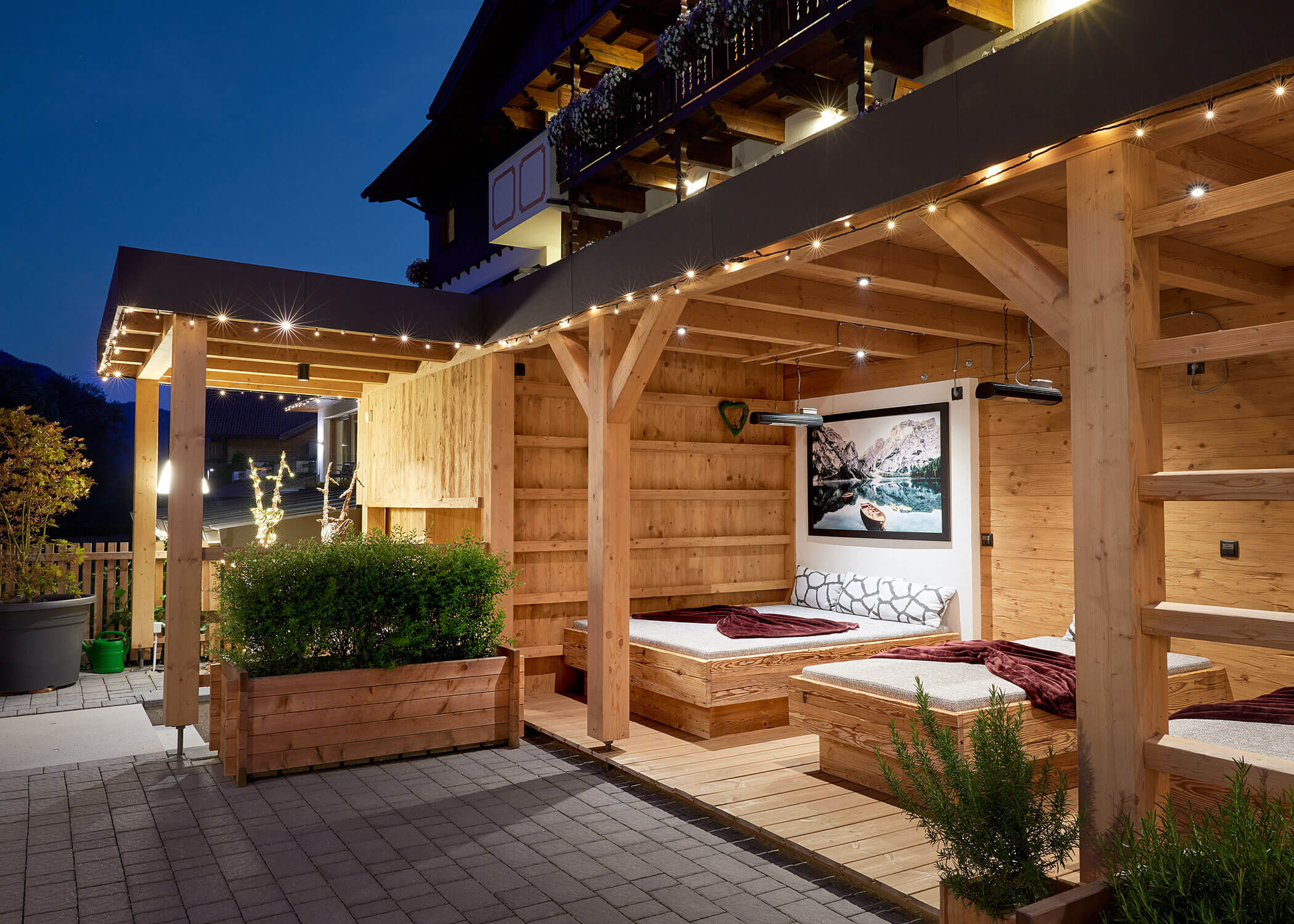 Wooden outdoor lounge area at night with cozy beds, string lights, and a mountain photo on the wall. - Hotel Magdalenahof
