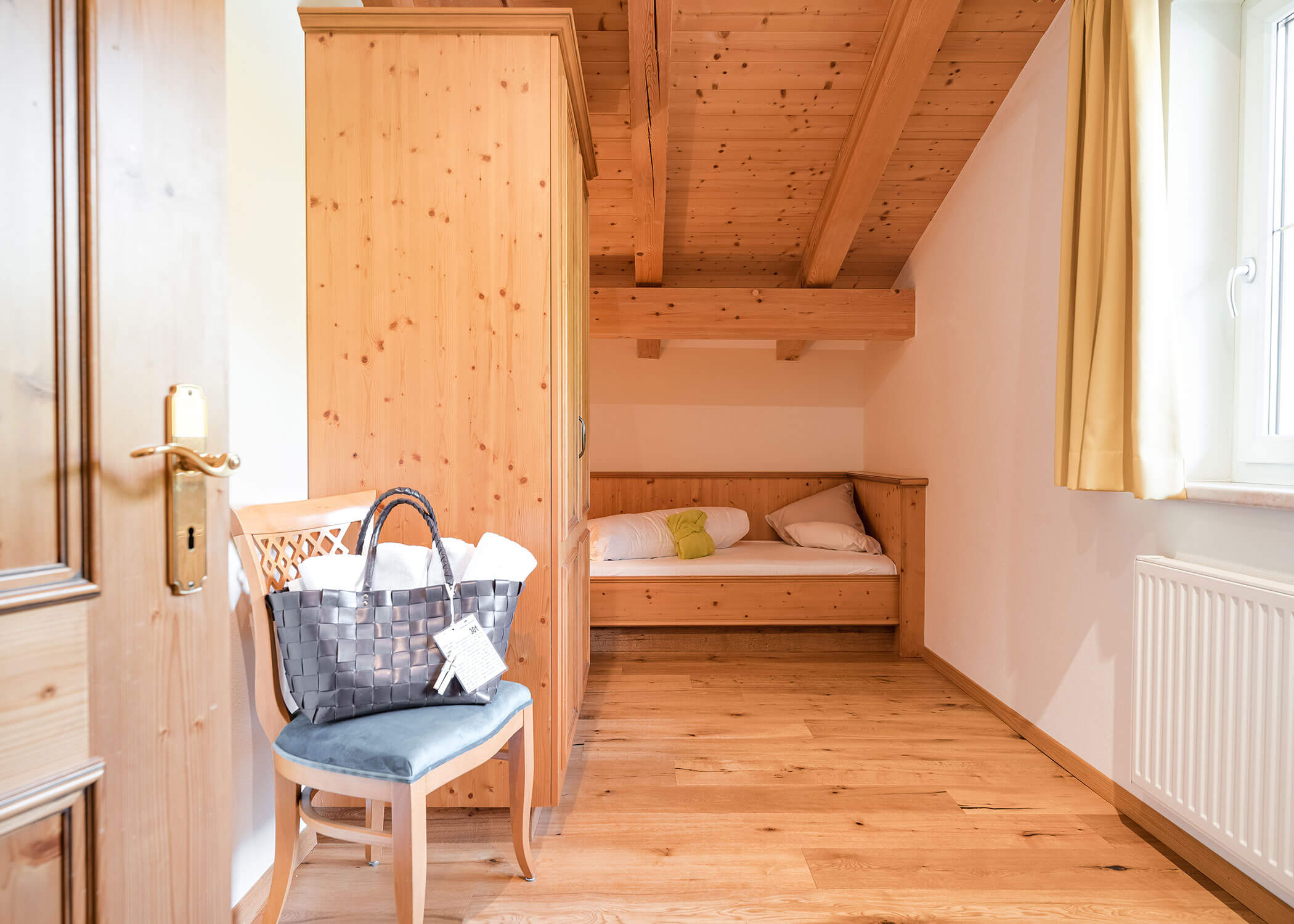 Gemütliches Holzzimmer mit Bett, Kleiderschrank, Fenster mit gelben Vorhängen und einer Tasche auf einem Stuhl. - Hotel Magdalenahof