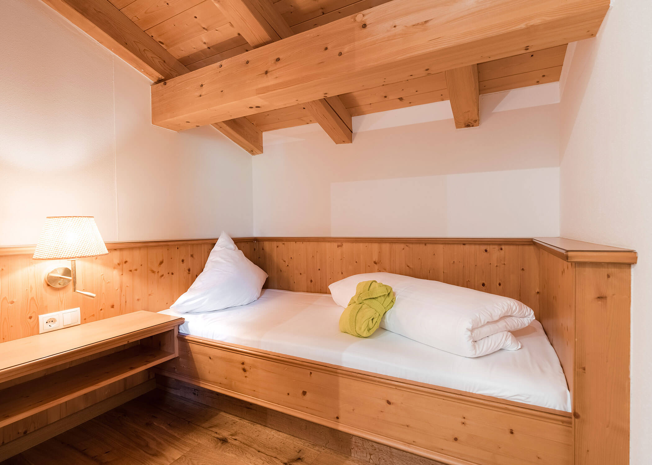 Gemütliches Holzzimmer mit einem Einzelbett, weißer Bettwäsche, einem grünen Handtuch und einer beleuchteten Wandlampe. - Hotel Magdalenahof