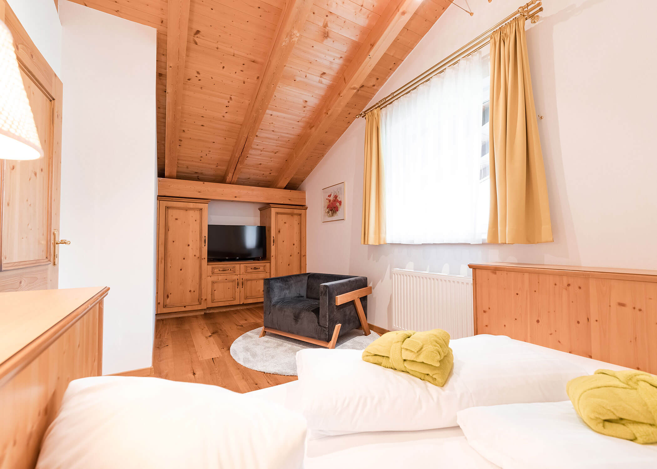 Gemütliches Schlafzimmer mit Holzdecke, TV, schwarzem Sessel und einem Bett mit grünen Handtüchern in der Nähe eines großen Fensters. - Hotel Magdalenahof
