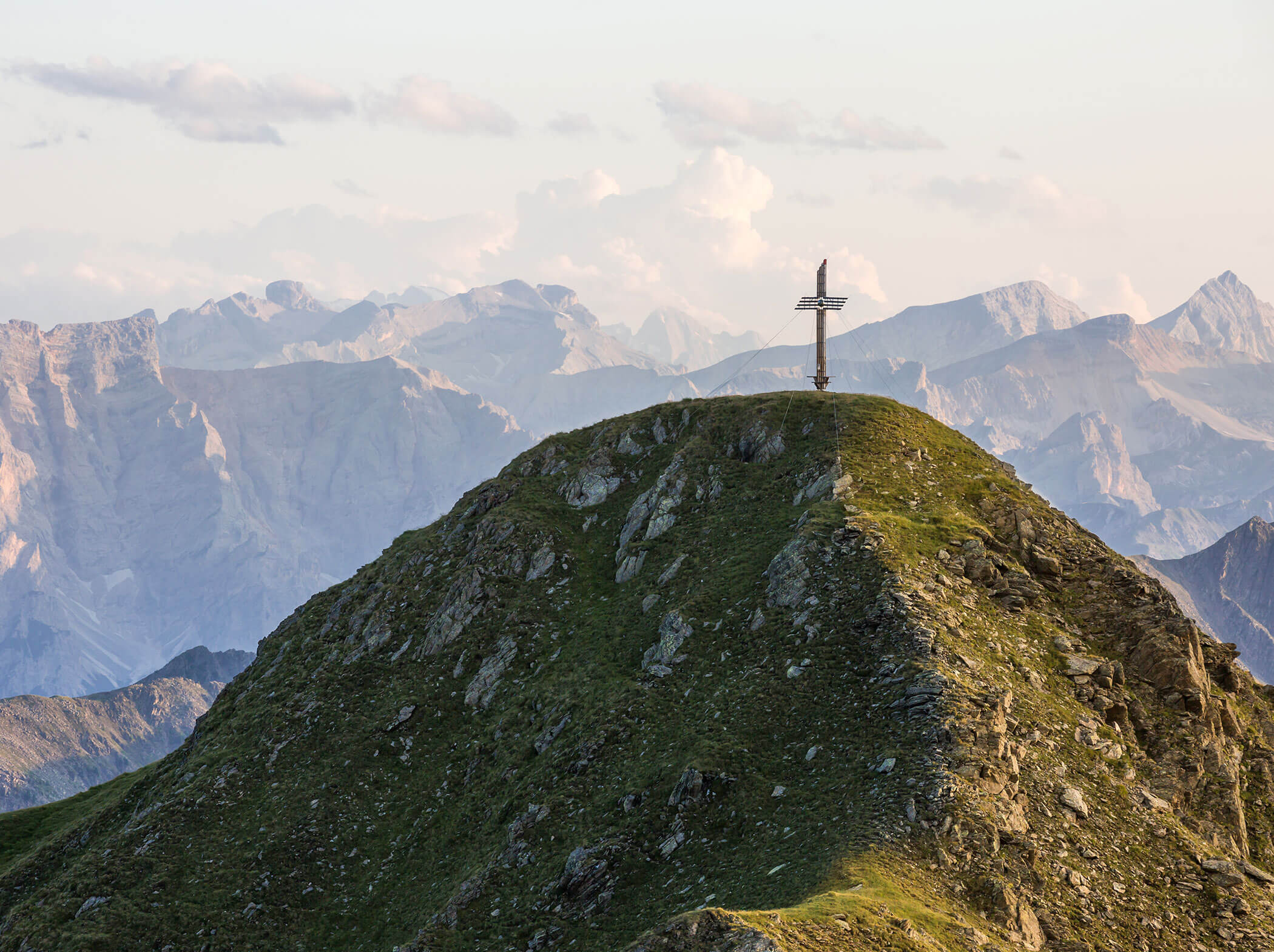 Ein Kreuz steht auf einem grasbewachsenen Berggipfel mit fernen, schroffen Bergen im Hintergrund. - Hotel Magdalenahof