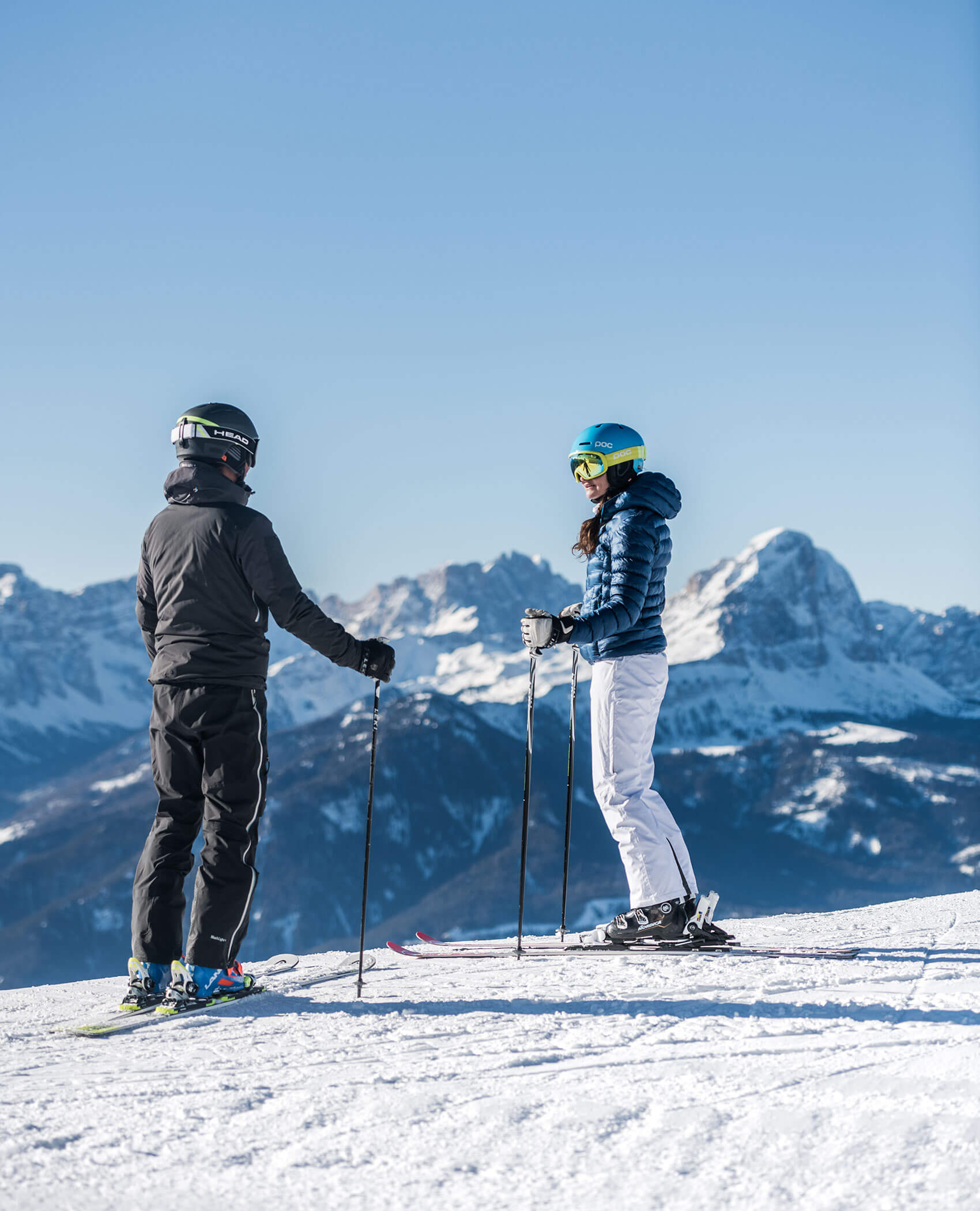Zwei Skifahrer stehen auf einer verschneiten Piste mit Skistöcken, Bergen und klarem blauen Himmel im Hintergrund. - Hotel Magdalenahof